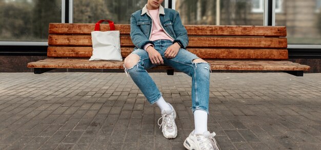 Trendiger junger Mann in stylischer Jeansjacke in lässigen Vintage-Jeans mit Stoffkäufer sitzt auf einer Holzbank an der Haltestelle des öffentlichen Gebäudes in der Stadt. Gut aussehender Kerl in modischer Kleidung auf der Straße.