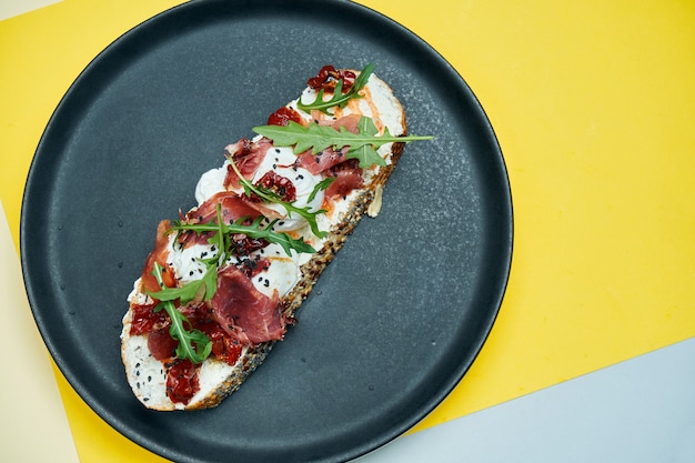 Trendiger Straßensnack. Leckeres Sandwich mit Jamon, pochierten Eiern, Gurke und Mikrogrün auf schwarzem Teller auf farbigem Oberflächenkopierraum.