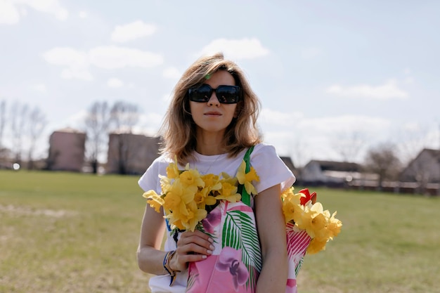 Trendiges Mädchen mit Kurzhaarfrisur in Sonnenbrille hält helle Tasche mit Blumen und posiert an sonnigen warmen Tagen auf natürlichem Hintergrund