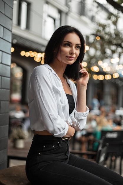 Trendiges, schönes Frauenmodell in modischer Kleidung mit weißem Hemd und schwarzer Jeans sitzt auf einem schwarzen Fass in der Stadt mit Bokeh-Lichtern