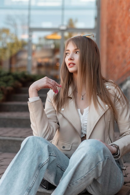 Trendiges, stilvolles, schönes junges Frauenmodell in modischer Freizeitkleidung mit Lederjacke und Vintage-Jeans sitzt auf den Stufen in der Nähe eines modernen Einkaufszentrums aus rotem Backstein