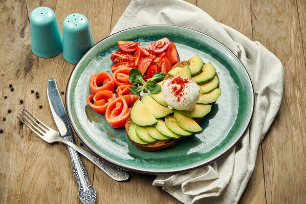 Trendnahrung - Avocado-Toast mit pochiertem Ei auf Roggenbrot mit seitlichem Lachs, Tomate in einer Keramikplatte auf einem Holztisch. Gesundes Frühstück