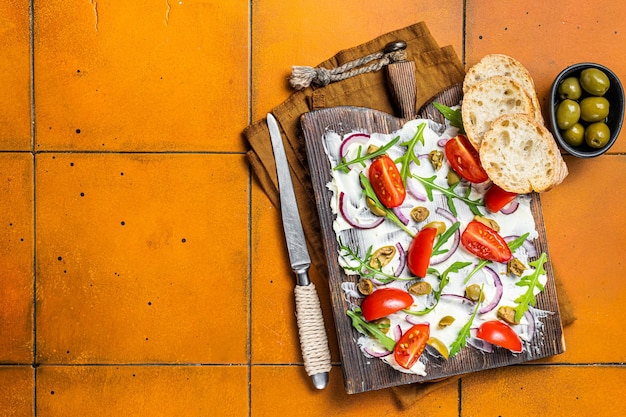 Trendy Butter Board mit Oliven, Zwiebeln, Rucola, Kapern und Tomaten Butterboard Orangefarbener Hintergrund Draufsicht Kopierbereich