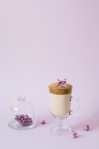 Trendy drink 2020 dalgon Café em um copo transparente com um tubo de papel, um prato com flores lilás em um fundo lilás com um espaço de cópia