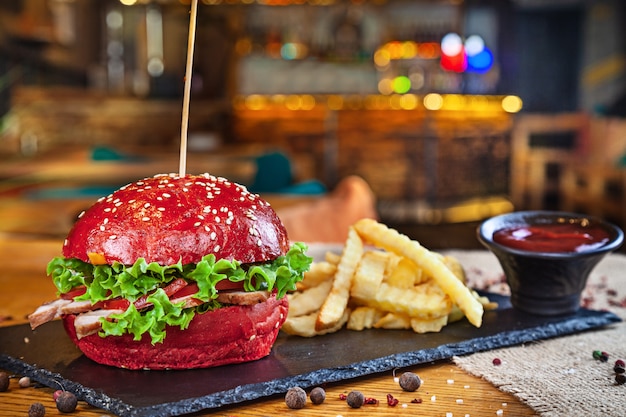 Trendy Red Bun Burger mit Schweinefleisch serviert mit Pommes Frites und roter Sauce. Ungesundes Fast Food. Burger im Restaurant Interieur. Amerikanischer Burger auf Steinplatte und Dachboden bacground mit Kopierraum. Sandwich