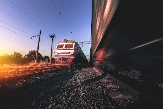 Trenes de carga y pasajeros en vías con efecto de desenfoque de movimiento al atardecer. Estación de tren en Ucrania