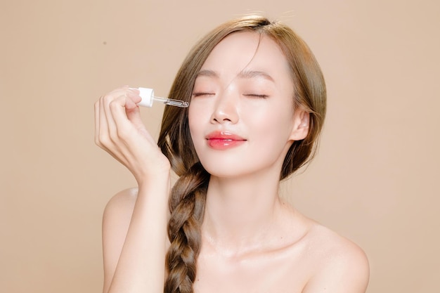 Trenza de cabello largo de mujer asiática joven con maquillaje natural en la cara y piel limpia y fresca con gotero para aplicar suero sobre fondo beige aislado Modelo femenino en estudio Tratamiento facial Cosmetología