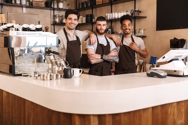 Três alegres baristas em frente ao balcão da cafeteria dentro de casa