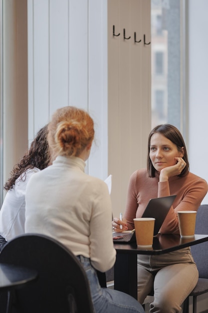 Foto três amigas e colegas em uma cafeteria estão discutindo um trabalho ou um projeto com tecnologias modernas de laptop