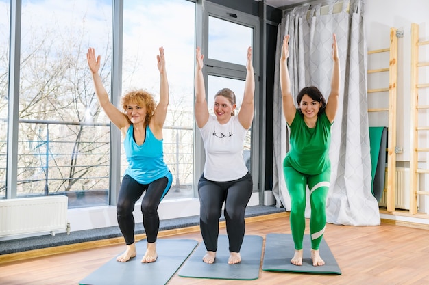 Três amigas fazem ioga pilates fitness juntas, treino em grupo, agachamento com as mãos para cima