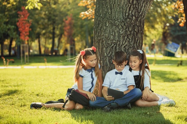 Tres amigos adolescentes escolares sonrientes sentados en el parque en el césped y jugar nuevo juego de tableta juntos. Diferentes emociones en sus caras