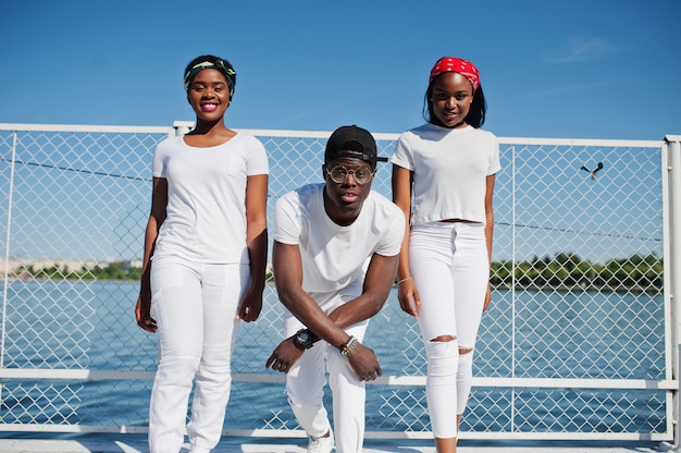 Três amigos afro-americanos à moda, vista-se na roupa branca no cais na praia contra a gaiola. Moda de rua dos jovens negros. Homem negro com duas garotas africanas.