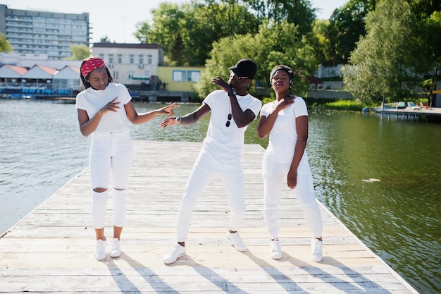 Três amigos afro-americanos elegantes usam roupas brancas no cais na praia mostrando moda de rua de jovens negros Homem negro com duas garotas africanas