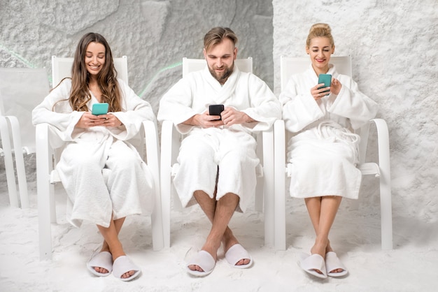 Tres amigos en batas de baño sentados con teléfonos, aplicando terapia de sal en la sala de sal
