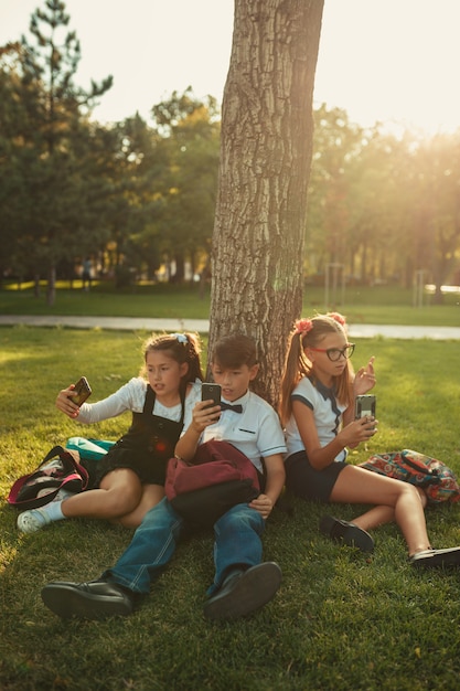 Tres amigos de la escuela están sentados en el césped debajo de un árbol y jugando con sus artilugios. Los amigos no se comunican en la vida prefiriendo el teléfono.