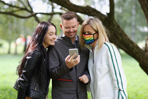 Tres amigos felices miran la pantalla del teléfono inteligente, una mujer lleva una máscara de arco iris