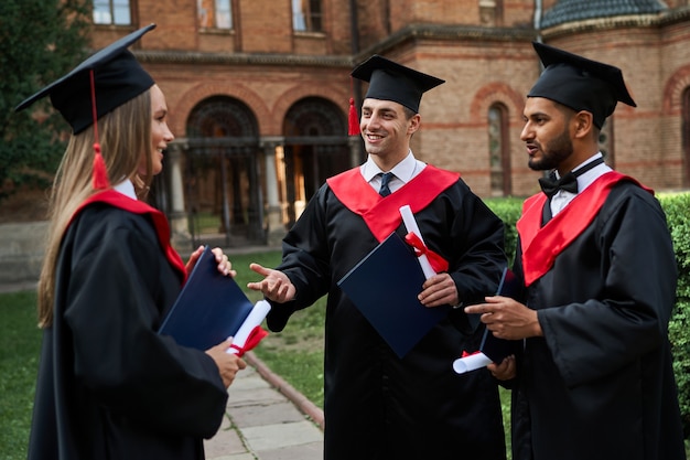 Tres amigos graduados en batas de graduación hablando en el campus con diploma en sus manos.