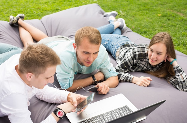 Tres amigos jovenes sonrientes felices que ponen en un amortiguador al aire libre y que miran la computadora portátil.