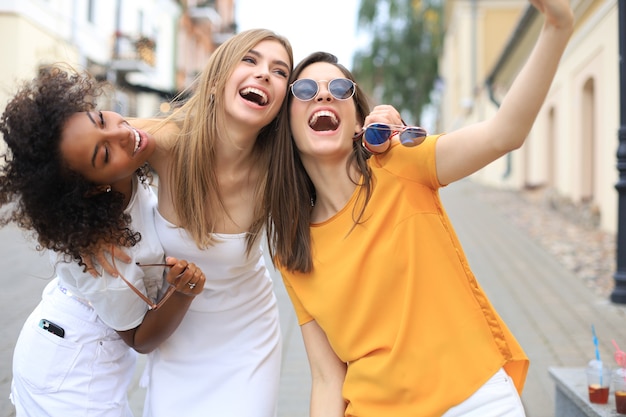 Três amigos lindos garotas se divertindo juntos, tirando uma selfie na cidade.