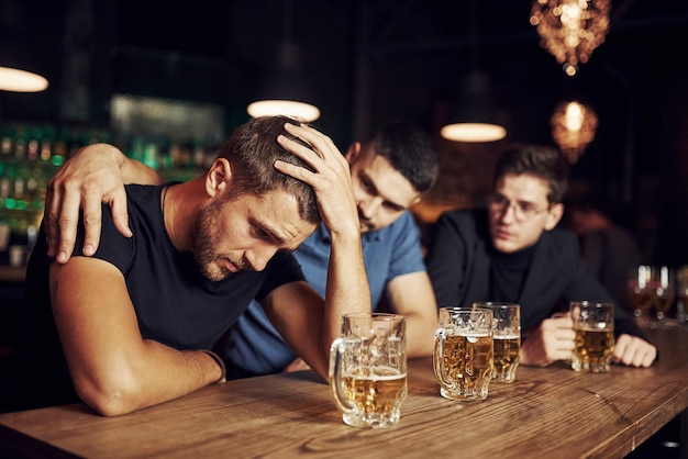 Tres amigos masculinos en el bar apoyando al triste amigo unidad de la gente con cerveza en la mesa