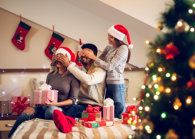 Três amigos muito brincalhões com chapéu de Papai Noel sentado na cama para as férias de Natal e fazendo um trem com os olhos fechados para um presente surpresa.