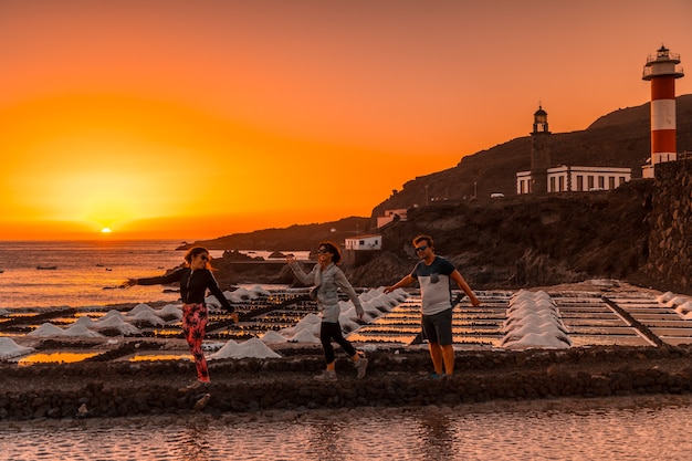 Três amigos no pôr do sol laranja nas salinas e ao fundo o Farol de Fuencaliente na rota dos vulcões ao sul da ilha de La Palma, nas Ilhas Canárias, Espanha