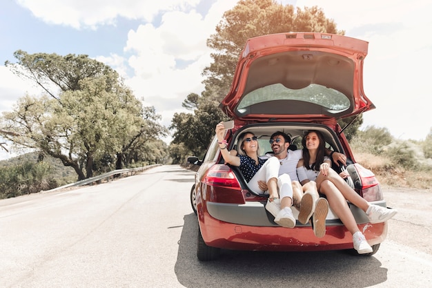 Foto tres amigos sentados juntos en el baúl del auto tomando autorretrato en la carretera
