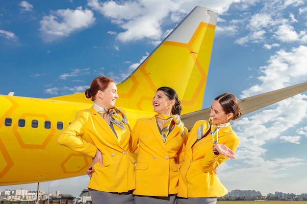Tres auxiliares de vuelo sonrientes con uniforme de línea aérea parados cerca de un avión amarillo contra el cielo nublado
