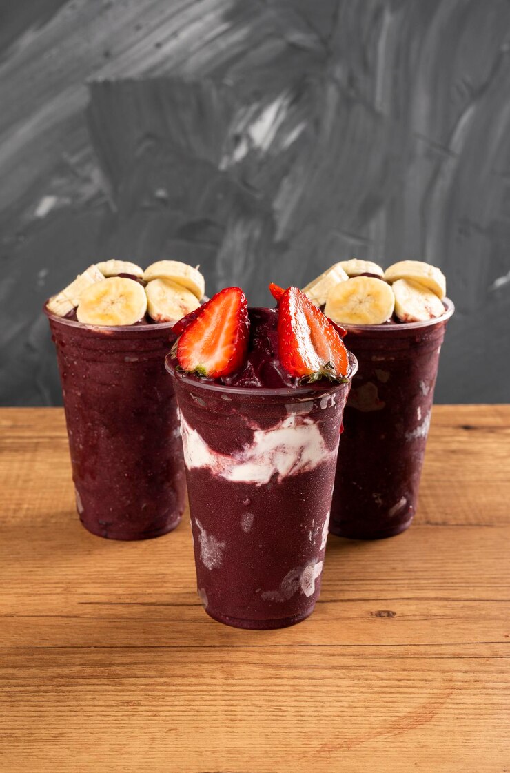 Foto três batidos de sorvete brasileiro de açaí berry em copo plástico com morangos, bananas e leite condensado. sobre uma mesa de madeira e um fundo cinza de verão. vista frontal para menu e mídia social