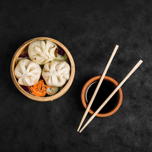 Tres bolas de masa hervida con ensalada en vaporeras y tazón de salsa de soja con palillos sobre fondo de textura negra