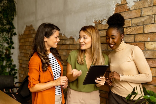 Três bonitas mulheres de negócios jovens com tablet digital de pé junto à parede de tijolos no escritório de estilo industrial