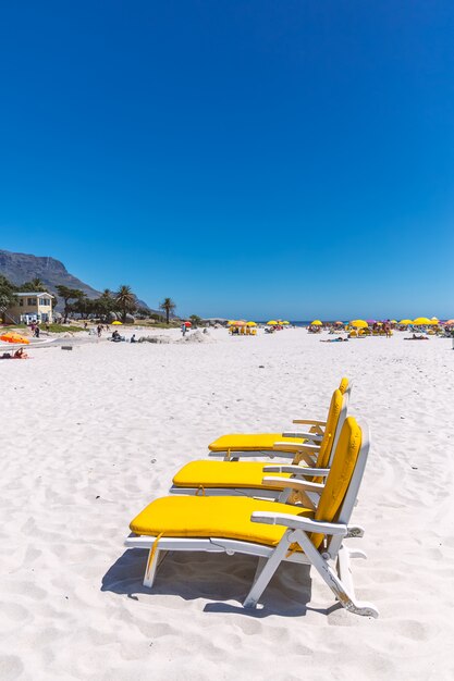 Três cadeiras de praia amarela em um dia ensolarado na praia de areia de camps bay, na cidade do cabo