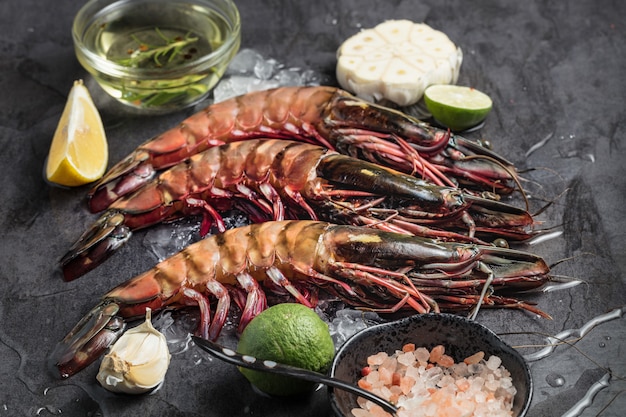 Três camarões enormes crus no gelo com ingredientes básicos para cozinhar em fundo escuro.