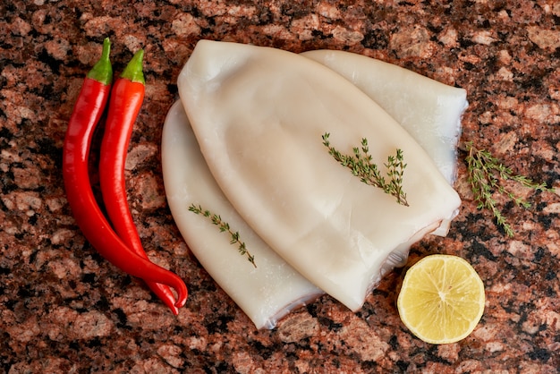 Tres canales de calamares crudos frescos con limón, pimienta, tomillo sobre una mesa oscura de piedra de mármol.