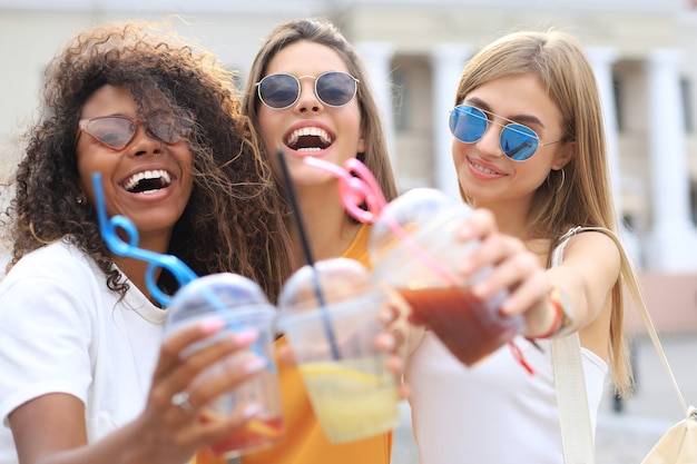Tres chicas hipster cool de moda, amigos beben cócteles en el fondo de la ciudad urbana.
