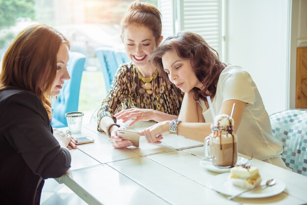 Tres chicas lindas viendo algo divertido en el teléfono inteligente en el café. Señora bonita morena sosteniendo el teléfono celular en sus manos