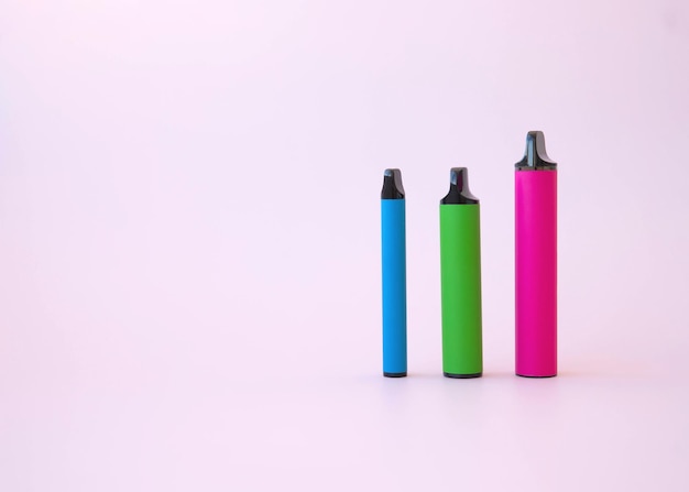 Tres cigarrillos electrónicos rosa brillante verde y azul de diferentes tamaños sobre un fondo rosa