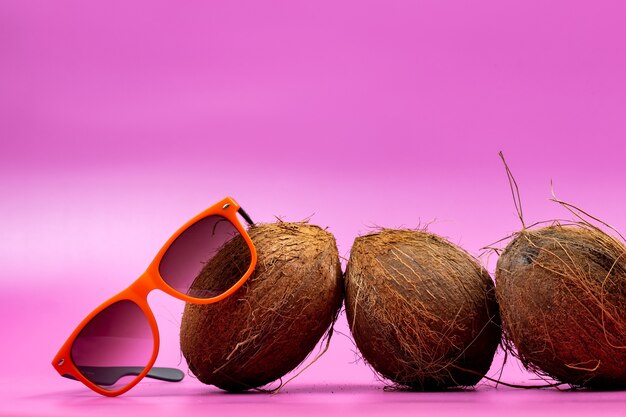 Tres cocos enteros y vasos naranjas sobre un fondo rosa.