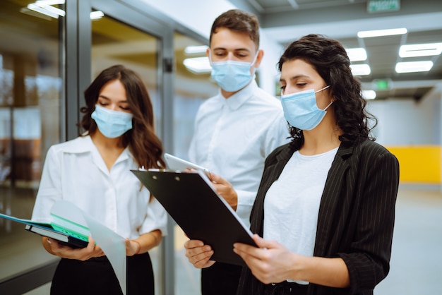 Tres colegas de negocios en mascarilla protectora discutiendo asuntos relacionados con el trabajo en una oficina.