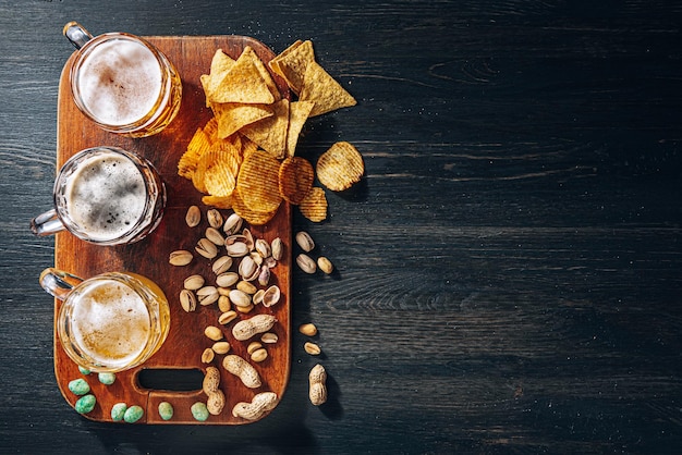 Três copos de cerveja artesanal cara, clássica e sem filtro e escura em um copo sobre a mesa com um lanche de chips de amendoim e pistache e nachos