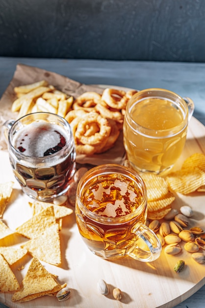 Três copos de cerveja artesanal cara, clássicos e não filtrados e escuros em um copo em cima da mesa com um lanche de batatas e nachos de amendoim e pistache