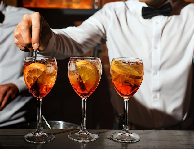Três copos de vidro com um coquetel de aperol spritz estão no bar, o barman está mexendo um dos aperitivos