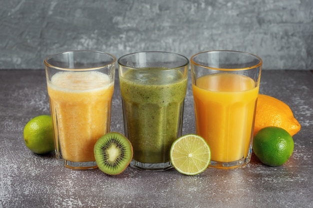 Três copos seguidos com suco de laranja e banana, kiwi, laranja, batido de espinafre, rodeado por metades de frutas em um fundo cinza de concreto. o conceito de perder peso e alimentação adequada.