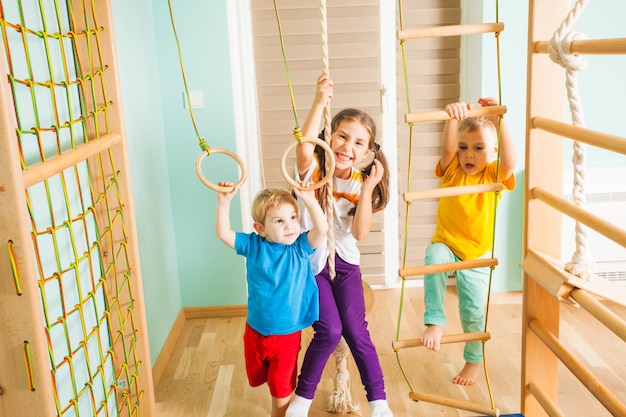 Três crianças ativas em camisetas coloridas, exercitando juntas, penduradas e subindo em uma corda, escada e anéis em seu complexo esportivo. Conceito de estilo de vida familiar ativo