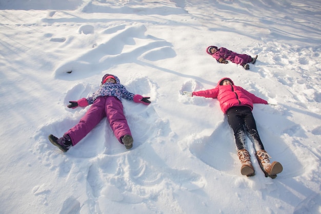 Três crianças fazem anjos de neve em um dia ensolarado de inverno