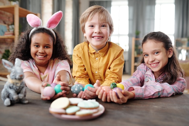 Três crianças interculturais felizes em trajes casuais segurando ovos de Páscoa pintados e olhando para você com sorrisos cheios de dentes enquanto estão sentados à mesa