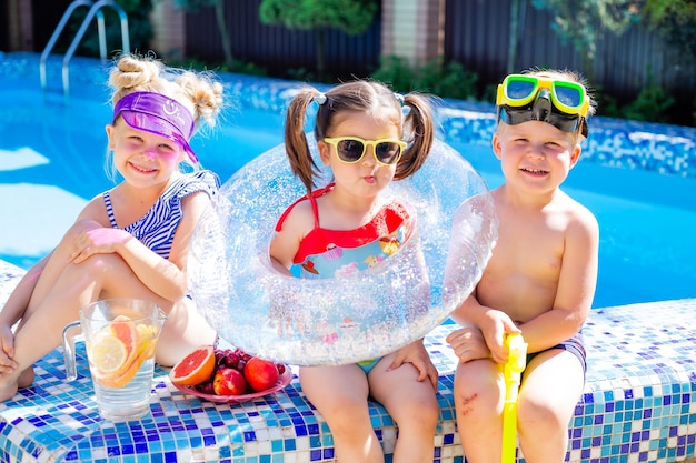 Foto três crianças no verão sentam-se à beira da piscina de óculos escuros e bebem limonada