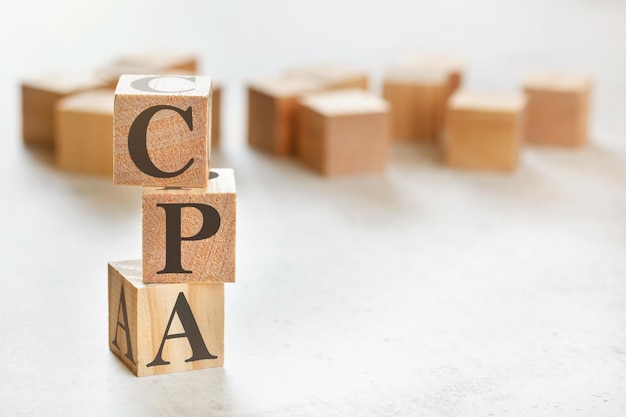 Três cubos de madeira com letras CPA (significa Custo por ação/aquisição), sobre mesa branca, mais ao fundo, espaço para texto no canto inferior direito