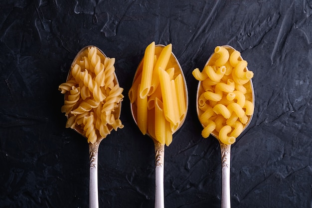 Tres cucharas de cubiertos con variedad de pasta de trigo dorado sin cocer en una mesa con textura negra oscura, vista superior