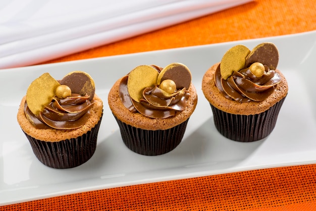 Tres cupcakes de chocolate con detalles dorados sobre vajilla blanca y fondo naranja.